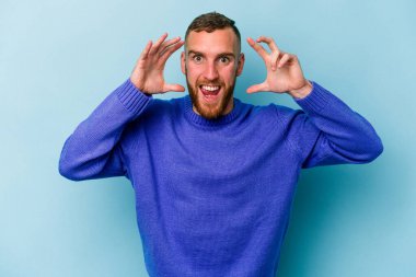 Mavi arka planda izole edilmiş genç beyaz adam hoş bir sürpriz alıyor, heyecanlı ve el kaldırıyor..