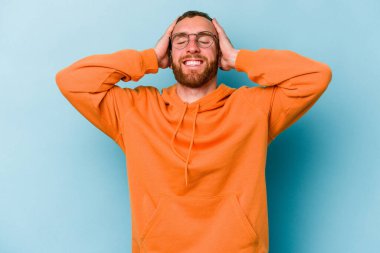 Mavi arka planda izole edilmiş genç beyaz adam ellerini başının üstünde tutarak neşe içinde gülüyor. Mutluluk kavramı.