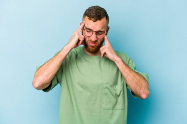 Genç beyaz adam mavi arka planda izole edilmiş tapınaklara dokunuyor ve başı ağrıyor..