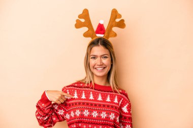 Young caucasian woman wearing a christmas reindeer hat isolated on beige background person pointing by hand to a shirt copy space, proud and confident
