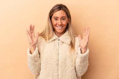 Beyaz tenli genç bir kadın, bej arka planda izole edilmiş. Yüksek sesle gülüyor, elini göğsüne koyuyor..