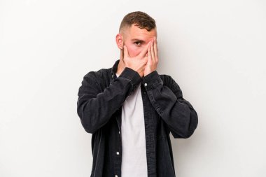 Beyaz arka planda izole edilmiş genç beyaz bir adam parmaklarının arasından korkmuş ve gergin bir şekilde göz kırpıyor..