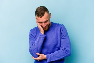 Mavi arka planda izole edilmiş genç beyaz adam mutlu bir şekilde gülüyor ve ellerini karnının üstünde tutarak eğleniyor..