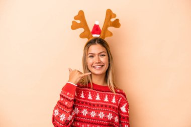 Young caucasian woman wearing a christmas reindeer hat isolated on beige background showing a mobile phone call gesture with fingers.