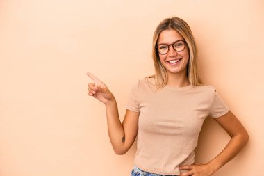 Beyaz tenli genç bir kadın, bej arka planda izole edilmiş. Gülümsüyor, işaret parmağıyla işaret ediyor..