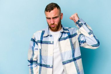 Mavi arka planda izole edilmiş genç beyaz adam yumruğunu kameraya gösteriyor, agresif yüz ifadesi var..