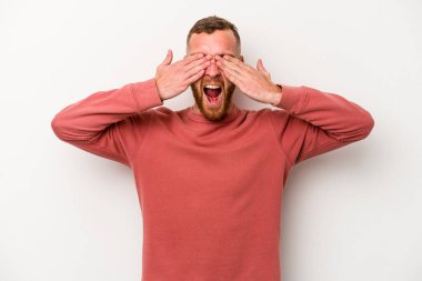 Beyaz arka planda soyutlanmış beyaz tenli genç bir adam elleriyle gözlerini kapatıyor, geniş bir gülümsemeyle bir sürpriz bekliyor..