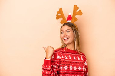 Young caucasian woman wearing a christmas reindeer hat isolated on beige background points with thumb finger away, laughing and carefree.