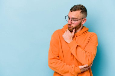 Mavi arka planda izole edilmiş genç beyaz adam şüpheli ve şüpheci bir ifadeyle yana bakıyor..