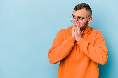 Kafkasyalı genç adam mavi arka planda izole edilmiş. Düşünceli bir şekilde ağzıyla ağzını kapatan bir fotokopi odasına bakıyor..