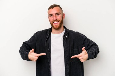 Beyaz arka planda yaşayan beyaz tenli genç bir adam gömlek fotokopi alanını gösteriyor, gururlu ve kendinden emin.