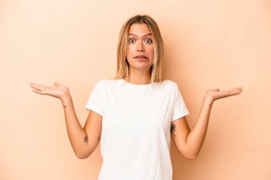 Beyaz tenli genç bir kadın, bej arka planda izole edilmiş. Kafası karışmış ve omuz silkiyor. Kopya alanını tutmak için..