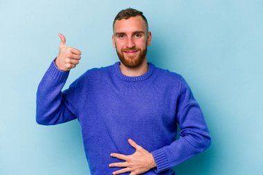 Mavi arka planda izole edilmiş genç beyaz adam karnına dokunuyor, nazikçe gülümsüyor, yemek yiyor ve tatmin oluyor..