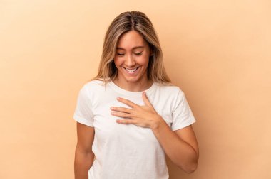 Beyaz tenli genç bir kadın, bej arka planda izole edilmiş. Yüksek sesle gülüyor, elini göğsüne koyuyor..