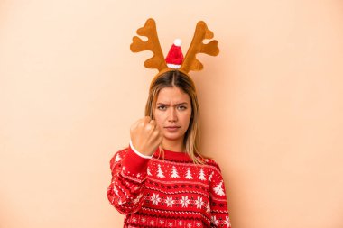 Young caucasian woman wearing a christmas reindeer hat isolated on beige background showing fist to camera, aggressive facial expression.