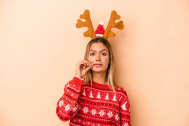 Young caucasian woman wearing a christmas reindeer hat isolated on beige background with fingers on lips keeping a secret.