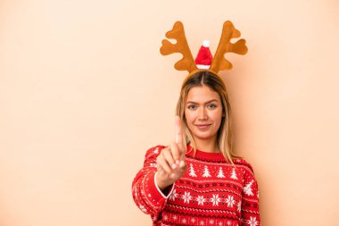 Young caucasian woman wearing a christmas reindeer hat isolated on beige background showing number one with finger.