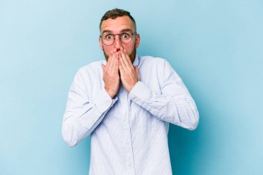 Mavi arka planda izole edilmiş genç beyaz adam elleriyle ağzını kapatırken şok oldu..