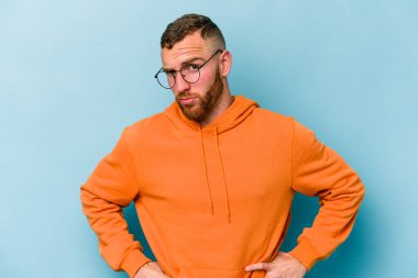 Kafkasyalı genç adam mavi arka planda izole edilmiş hoşnutsuzca kaşlarını çatmış, kollarını kavuşturmuş..