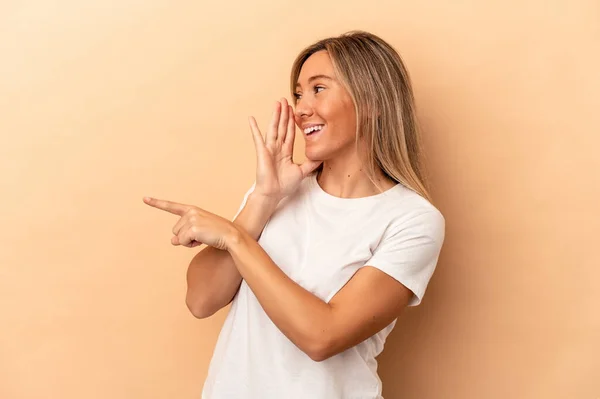 Young caucasian woman isolated on beige background saying a gossip, pointing to side reporting something.
