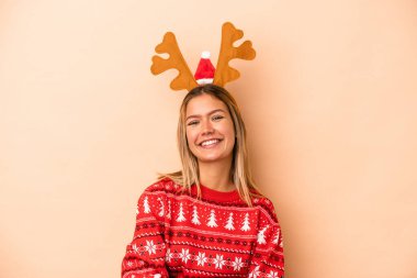 Young caucasian woman wearing a christmas reindeer hat isolated on beige background laughing and having fun.