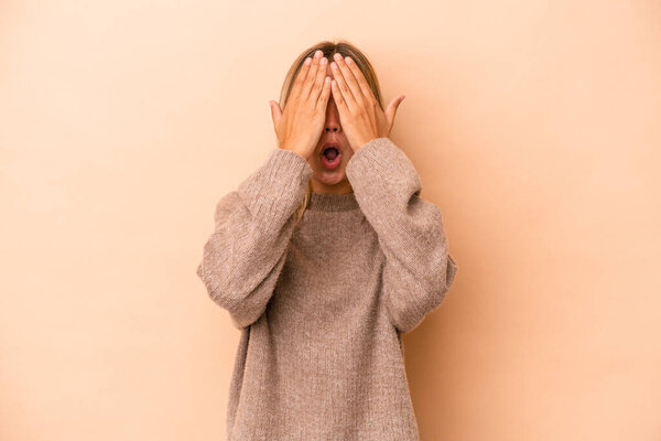 Молодая белая женщина, изолированная на бежевом фоне, весело прикрывающая половину лица ладонью.