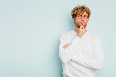 Mavi arka planda izole edilmiş genç beyaz adam şüpheli ve şüpheci bir ifadeyle yana bakıyor..