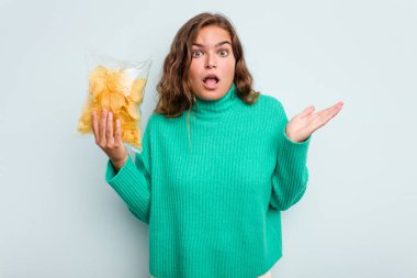 Mavi arka planda patates cipsi tutan beyaz kadın şaşırdı ve şok oldu..