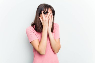 Beyaz arka planda izole edilmiş beyaz tenli genç bir kadın parmaklarının arasından korkmuş ve gergin bir şekilde göz kırpıyor..