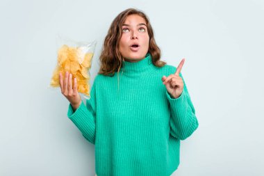 Elinde patates cipsi tutan beyaz bir kadın mavi arka planda izole edilmiş ve ağzı açık bir şekilde üst tarafı işaret ediyor..