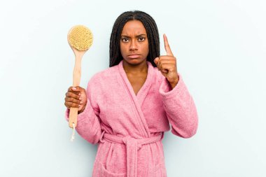 Elinde küvet fırçası olan genç Afrikalı bir kadın mavi arka planda izole edilmiş bir şekilde şakağı parmağıyla işaret ediyor, düşünüyor ve bir göreve odaklanıyor..