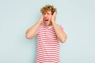 Kafkasyalı genç adam mavi arka planda izole edilmiş kulakları kulaklarıyla kapatıyor çok fazla ses duymamaya çalışıyor..