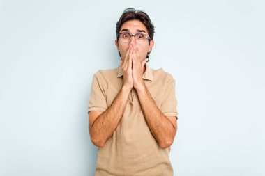 Genç İspanyol bir adam mavi arka planda izole edilmiş, ağzı kapalı ve elleri endişeli görünüyor..