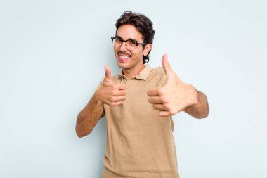 Genç İspanyol adam mavi arka planda izole edilmiş. Baş parmağını kaldırıyor, gülümsüyor ve kendinden emin..