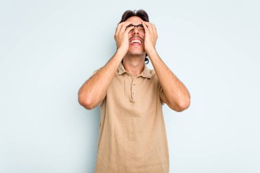 Mavi arka planda izole edilmiş genç İspanyol adam ellerini başının üstünde tutarak neşeyle gülüyor. Mutluluk kavramı.