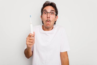 Beyaz arka planda izole edilmiş elektrikli diş fırçası tutan İspanyol genç adam omuz silkiyor ve gözleri açık..