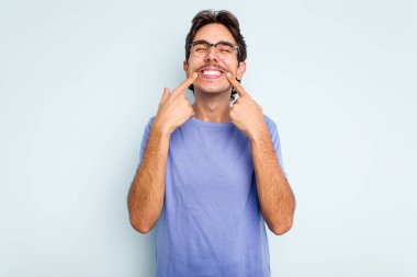 Mavi arka planda izole edilmiş genç İspanyol adam gülümsüyor, parmaklarını ağıza doğrultuyor..