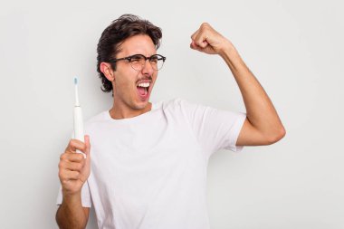 Beyaz arka planda elektrikli diş fırçası tutan İspanyol genç adam zafer kazandıktan sonra yumruğunu kaldırdı..