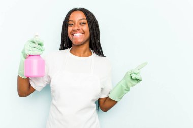 Genç Afrikalı Amerikalı temizlikçi kadın mavi arka planda izole edilmiş gülümsüyor ve boş alanda bir şey gösteriyor..