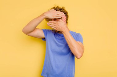 Sarı arka planda izole edilmiş genç beyaz adam kameraya parmaklarıyla göz kırpıyor, yüzünü kapatmaktan utanıyor..