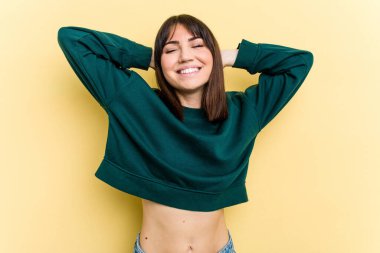 Sarı arka planda izole edilmiş genç beyaz bir kadın kendini güvende hissediyor, elleri başının arkasında..