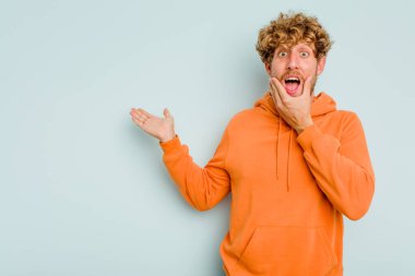 Mavi arka planda izole edilmiş genç beyaz bir adam avucunun içinde fotokopi alanı tutuyor, el üstünde tutuyor. Şaşırmış ve mutlu..