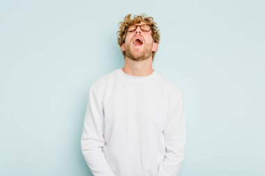 Kafkasyalı genç adam mavi arka planda izole edilmiş rahat ve mutlu bir şekilde gülüyor, boynunda dişlerini gösteriyor..