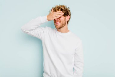 Mavi arka planda soyutlanmış genç beyaz bir adam elleriyle gözlerini kapatıyor, geniş bir gülümsemeyle bir sürpriz bekliyor..