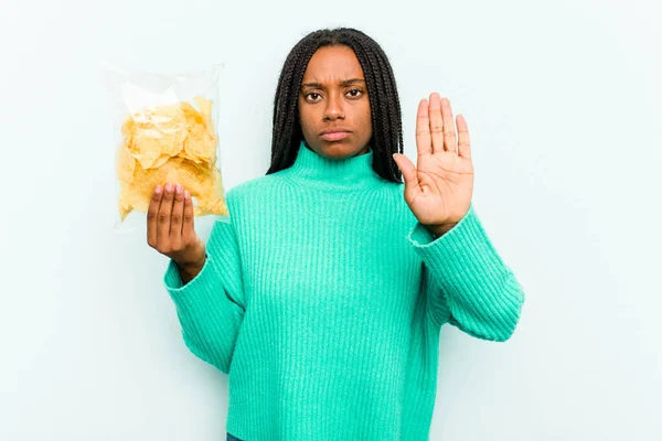 年轻的非洲裔美国女人拿着用蓝色背景隔开的薯片站在那里 伸出的手显示出停止的迹象 阻止了你 — 图库照片