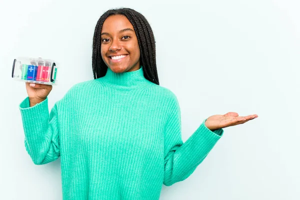 青い背景に隔離されたバッテリーボックスを持っている若いアフリカ系アメリカ人女性は 手のひらにコピースペースを示し 腰に別の手を保持します — ストック写真