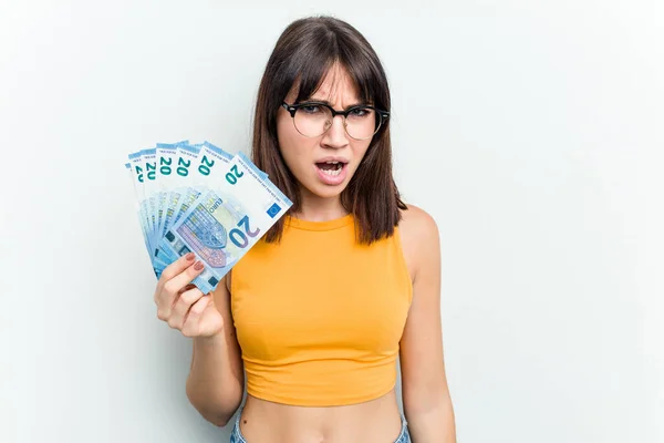 Young caucasian woman holding a banknotes isolated on blue background screaming very angry and aggressive.