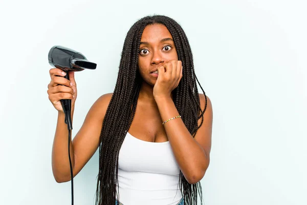 年轻的非洲裔美国女人拿着一个吹风机 用蓝色背景隔离 咬着指甲 神经紧张 非常焦虑 — 图库照片