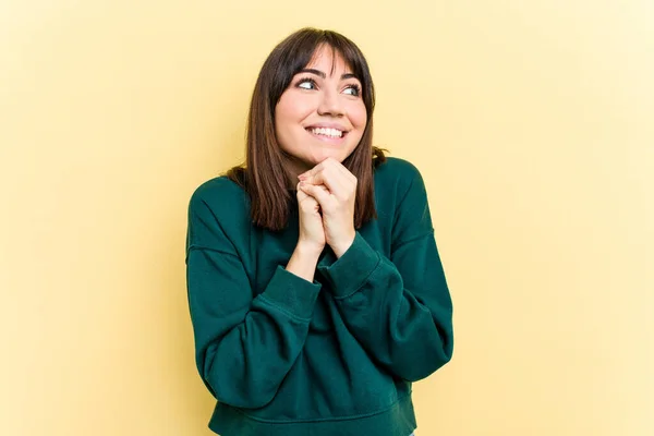 Young caucasian woman isolated on yellow background keeps hands under chin, is looking happily aside.