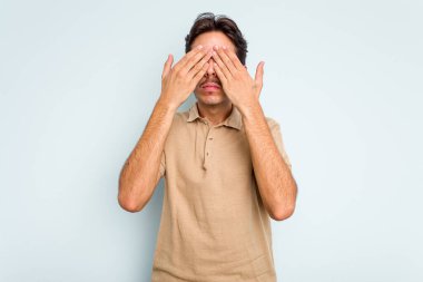 Mavi arka planda izole edilmiş genç İspanyol adam elleriyle gözlerini kapatmaktan korkuyor..